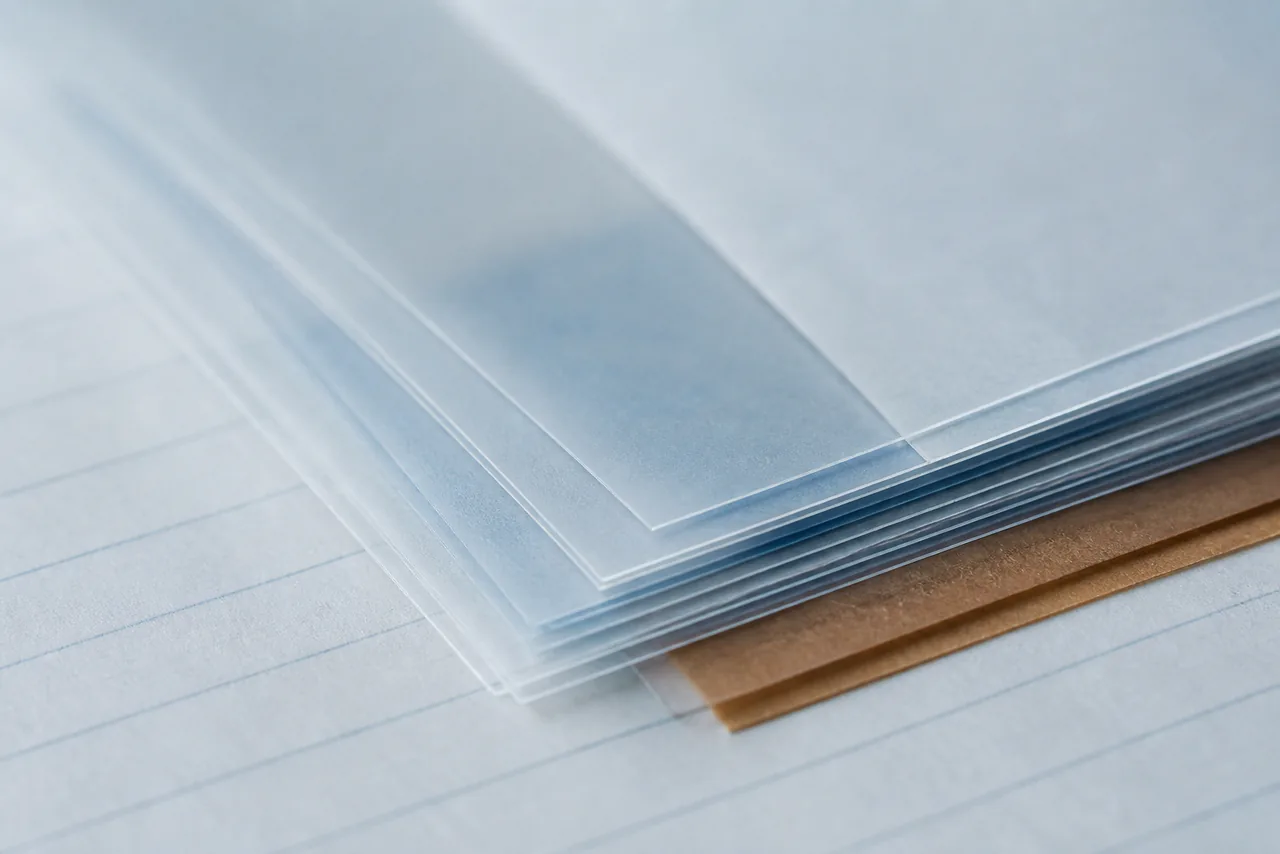 Translucent acetate sheets layered slightly out of register over pale blue ruled paper, their stacked edges forming fine ridges, with a single narrow warm brown paper strip showing beneath one corner.